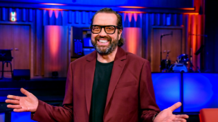 Moderator steht lächelnd auf einer bunt beleuchteten Studiobühne und breitet die Arme aus