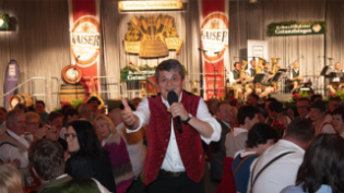 Moderator Josef "Bäff" Piendl steht vor dem Publikum und der Bühne des Kaltenhauser Gstanzlsingens. 