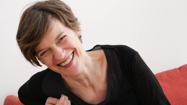 Lächelnde Frau mit kurzen Haaren sitzt auf einem Sofa.