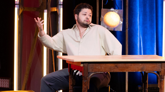 Mann sitzt auf einer Bühne an einem Tisch und gestikuliert mit der Hand.