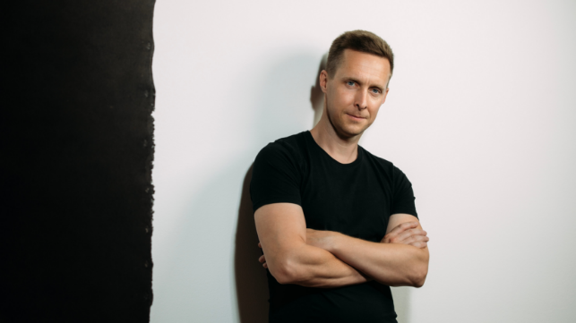 Mann mit verschränkten Armen steht vor einer hellen Wand.