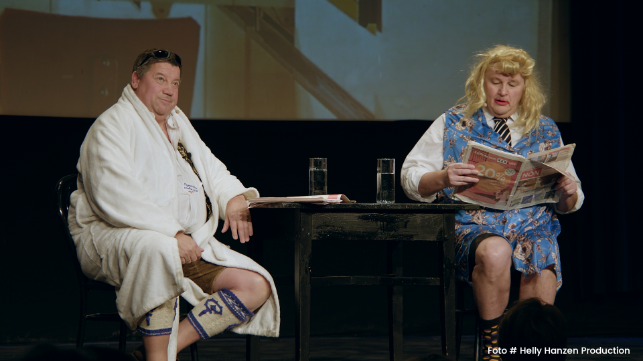 Zwei Personen sitzen auf einer Bühne an einem Tisch, einer liest Zeitung.