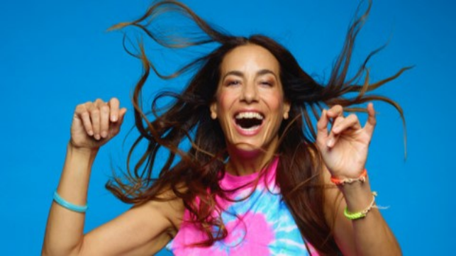 Frau mit langen Haaren lacht und tanzt vor blauem Hintergrund.