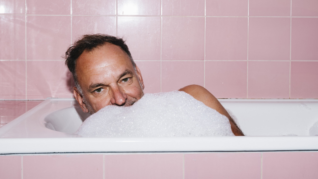 Mann liegt in einer Badewanne voller Schaum und schaut in die Kamera.