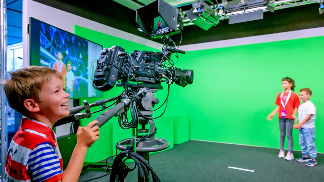 Blick in den Greenscreen. Ein kleiner Junge bedient die Kamera während zwei andere Kids lächelnd vor dem Greenscreen steht